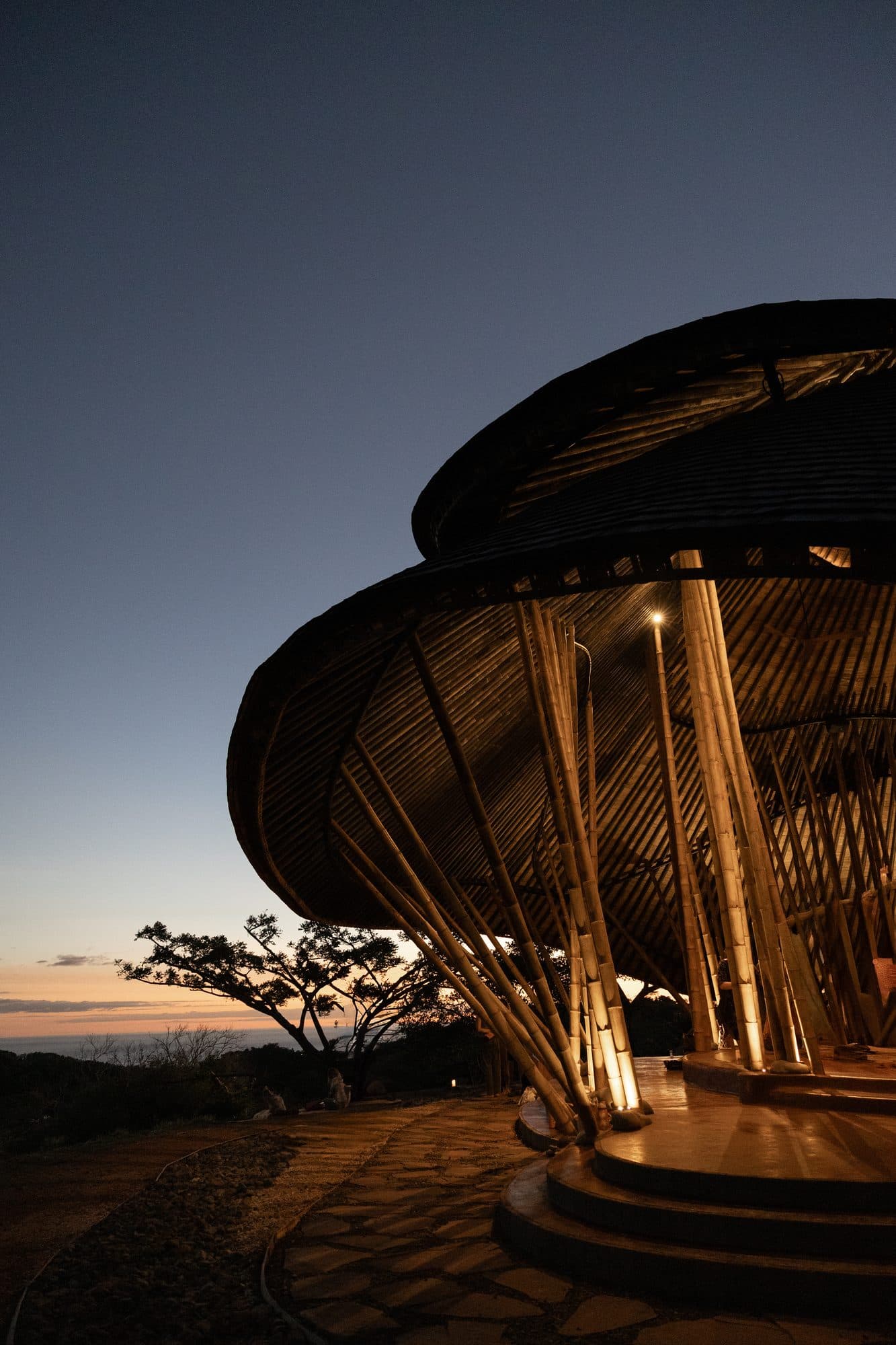 The bamboo maloca lit from within at dusk, the Pacific horizon behind it